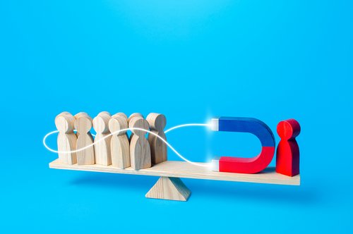 Waage mit Holzfiguren und eine rote Holzfigur die ein Magnet in der Hand hält Richtung der Gruppe