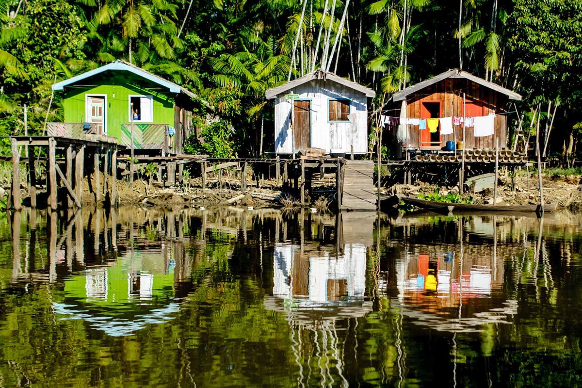 An einem Flussufer im Regendwald in Brasilien stehen drei Hütten aus Holz in unterschiedlichen Farben.