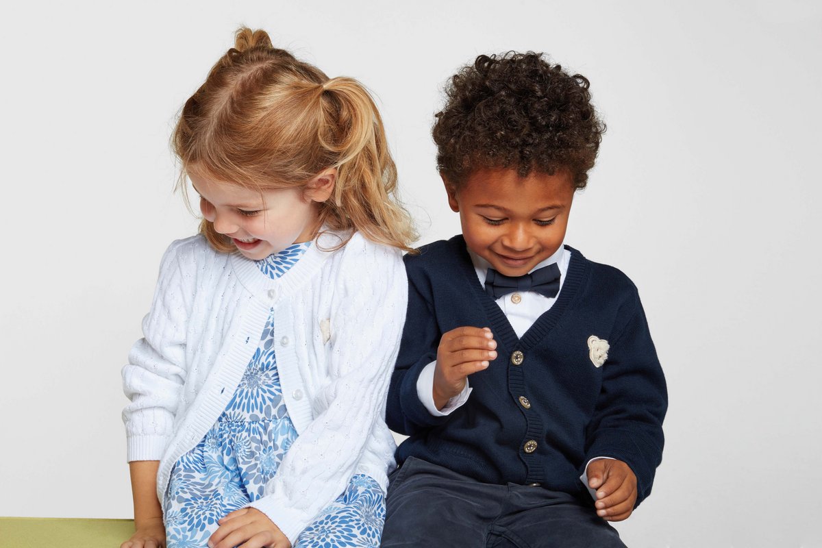 Ein Junge in einem Anzug von Steiff und ein Mädchen in einem hellblauen Kleid von Steiff sitzen nebeneinander und schauen an sich herunter.