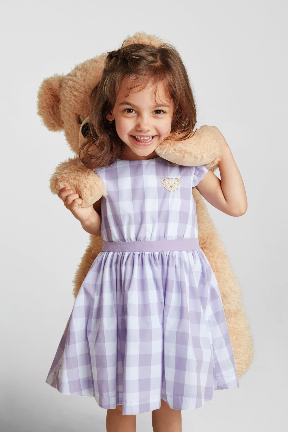Ein Mädchen in einem lila karierten Kleid trägt einen Steiff Teddybär auf dem Rücken.