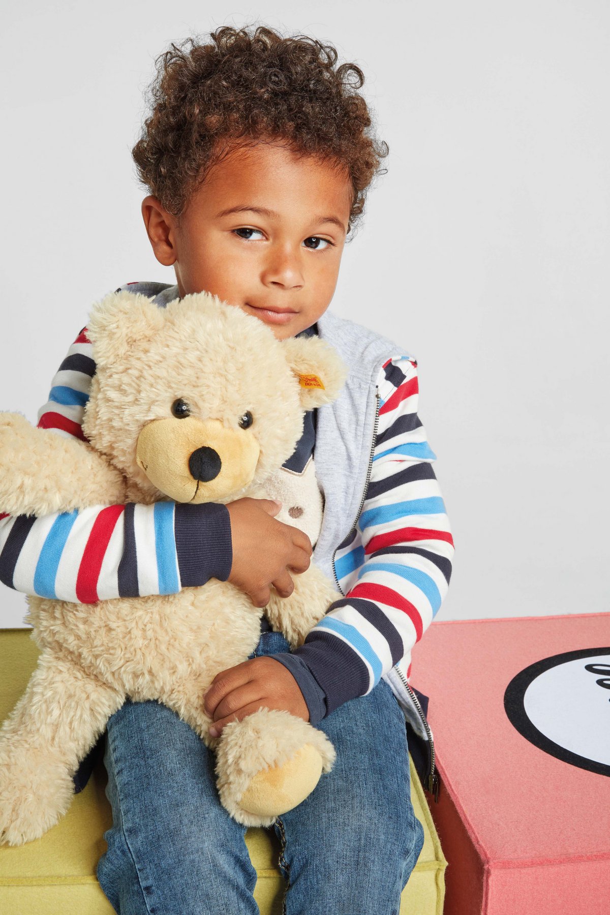 Ein Junge in einer gestreiften Jacke von Steiff sitzt mit einem Teddybär mit einem Kopf im Ohr in seinem Arm. 