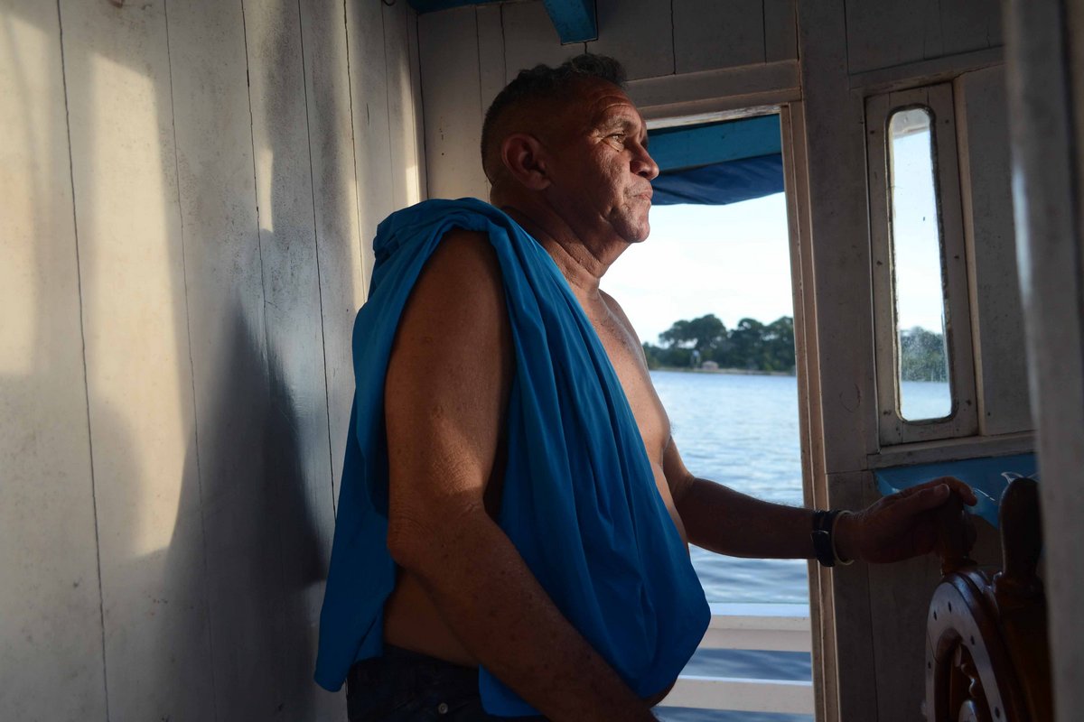 Ein Mann ohne T-Shirt steht mit einem blauen Tuch über der Schulter im Führerhäuschen eines Schiffs.