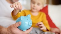 Kleinkind wirft ein Geldstück in ein blaues Porzellansparschwein, das von einem Erwachsenen gehalten wird.