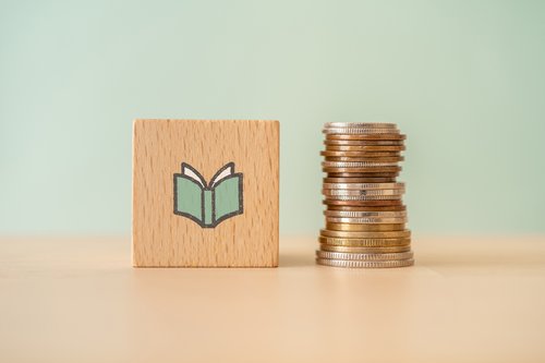 Neben einem Holzwürfel mit einem Buch darauf liegt ein Stapel Münzen.