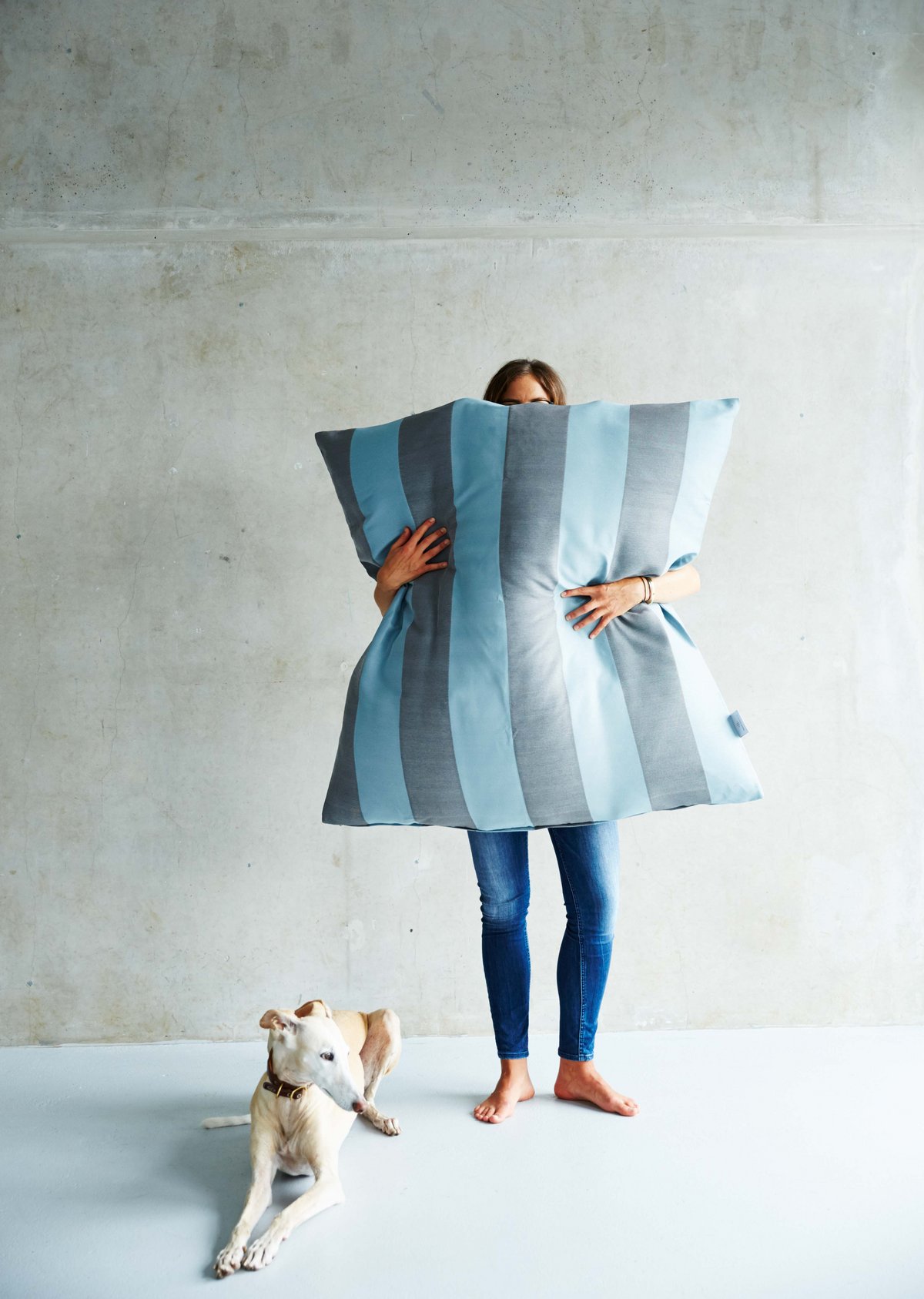 Eine Frau steht neben ihrem auf dem Boden liegenden Hund und versteckt sich hinter einem großen Hundekissen von Darling Little Place in blauen und grauen Streifen.
