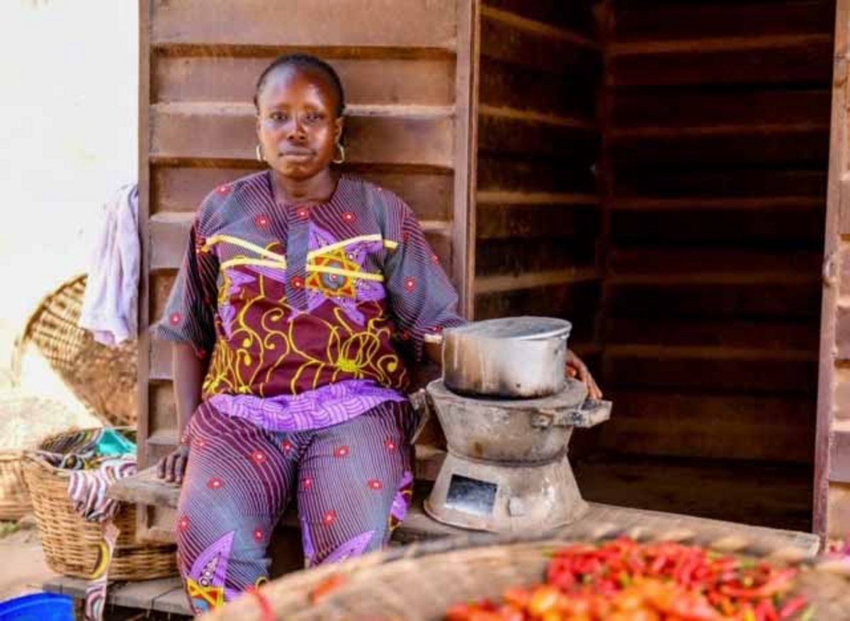 Eine nigerianische Frau lehnt in einem lila Gewand an der Wand eines Hauses, vor ihr steht ein Kochofen.