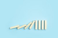 Vor hellblauem Hintergrund fallen helle Holzubausteine gegeneinander in einem Domino Effekt.