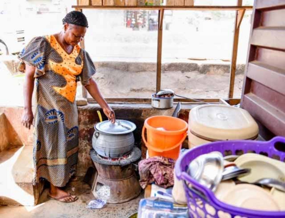 Vor einem Haus in Nigeria steht eine Frau vor mehreren Eimern und Töpfen und einem Kochofen.