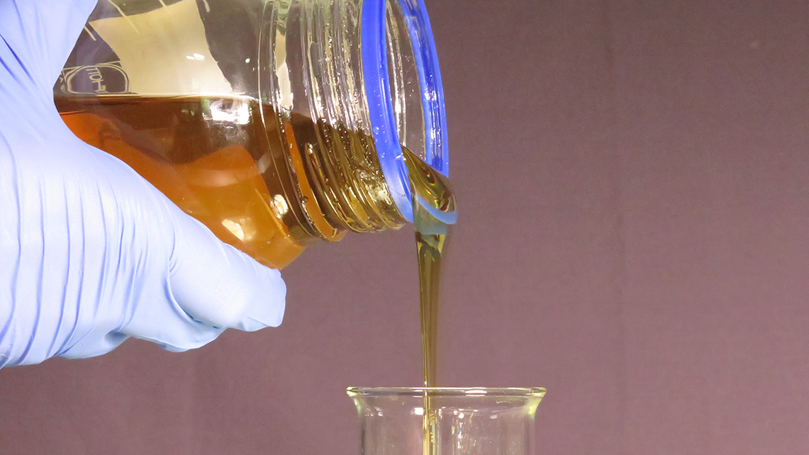 Behandschuhte Hand gießt bernsteinfarbene Flüssigkeit aus einem Schraubverschlussglas in einen Meßbecher