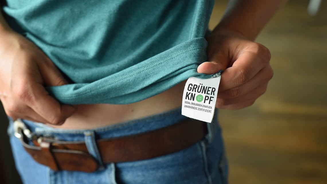 Frauenhand zeigt Innenlabel eines T-Shirts mit der Beschriftung Grüner Knopf