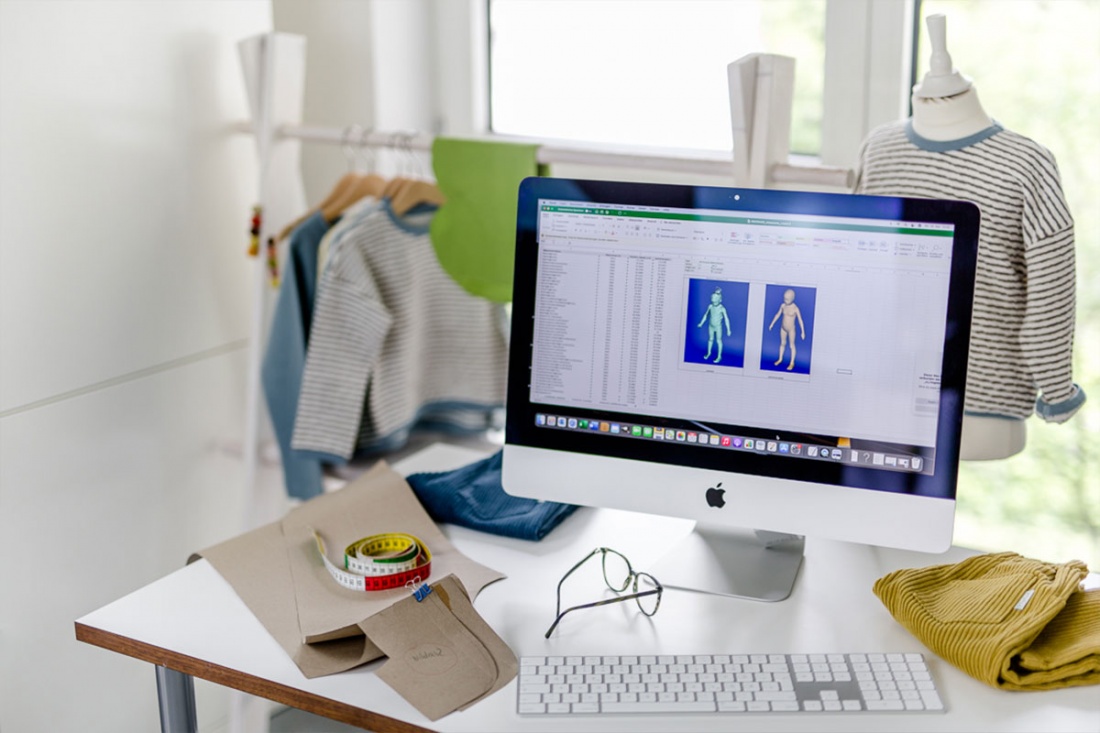 Computermonitor mit digitaler Erfassung von Körpermaßen steht auf einem Tisch neben verschiedenen Kleidungsstücken und Schnittmustern