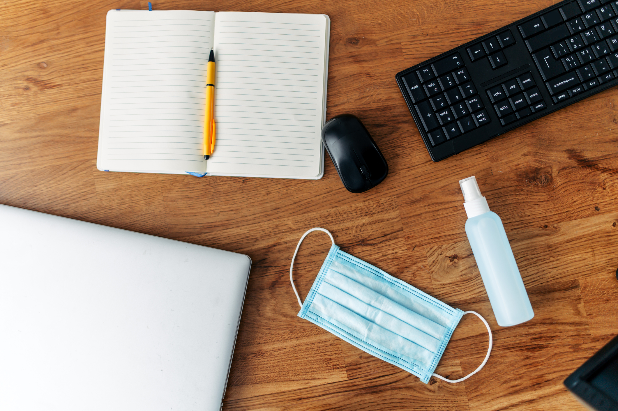 Auf einem Schreibtisch aus Holz liegen neben Laptop, Tastatur, Maus und einem offenen Notizbuch Desinfektionsspray und eine Maske.