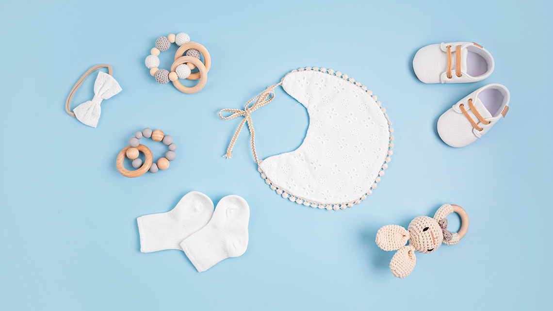 Auf hellblauem Hintergrund liegen verschiedene Babyartikel, darunter Schuhe, ein Lätzchen und eine Schleife.
