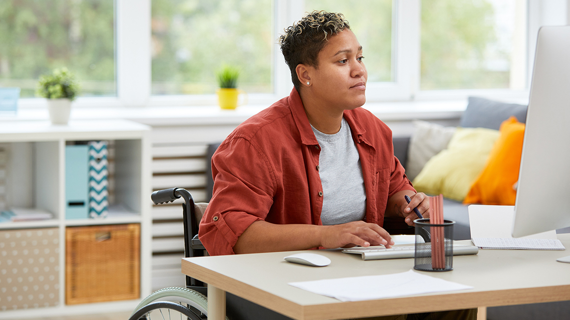 Frau in einem Rollstuhl sitzt an einem Schreibtisch und arbeitet an ihrem Computer.