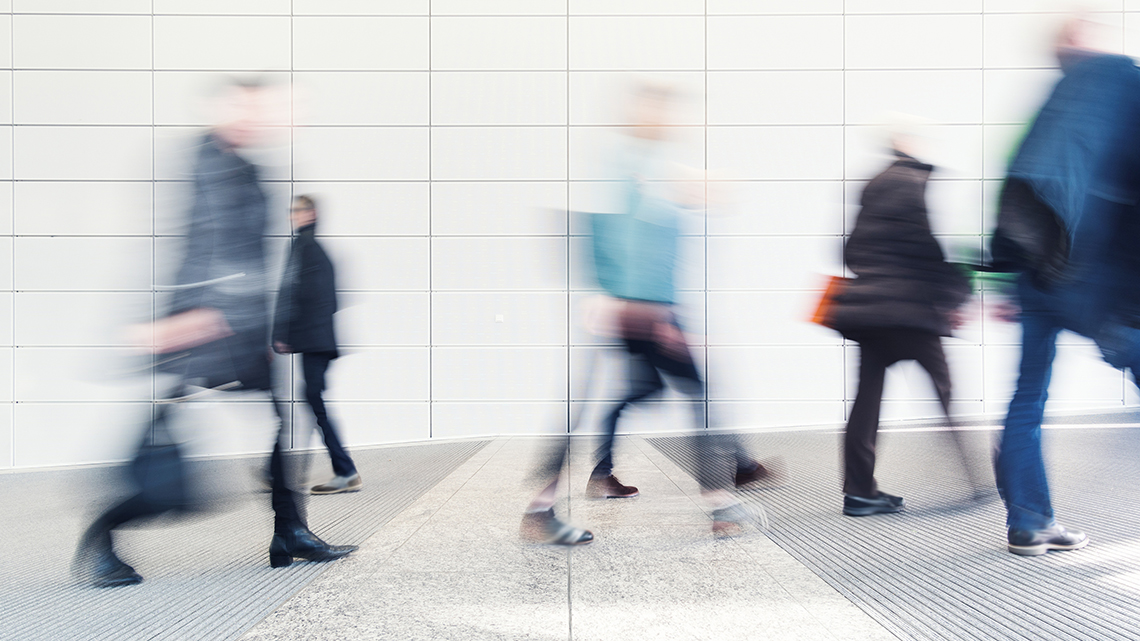 Viele Personen in geschäftlicher Kleidung bewegen sich vor einer Wand. 