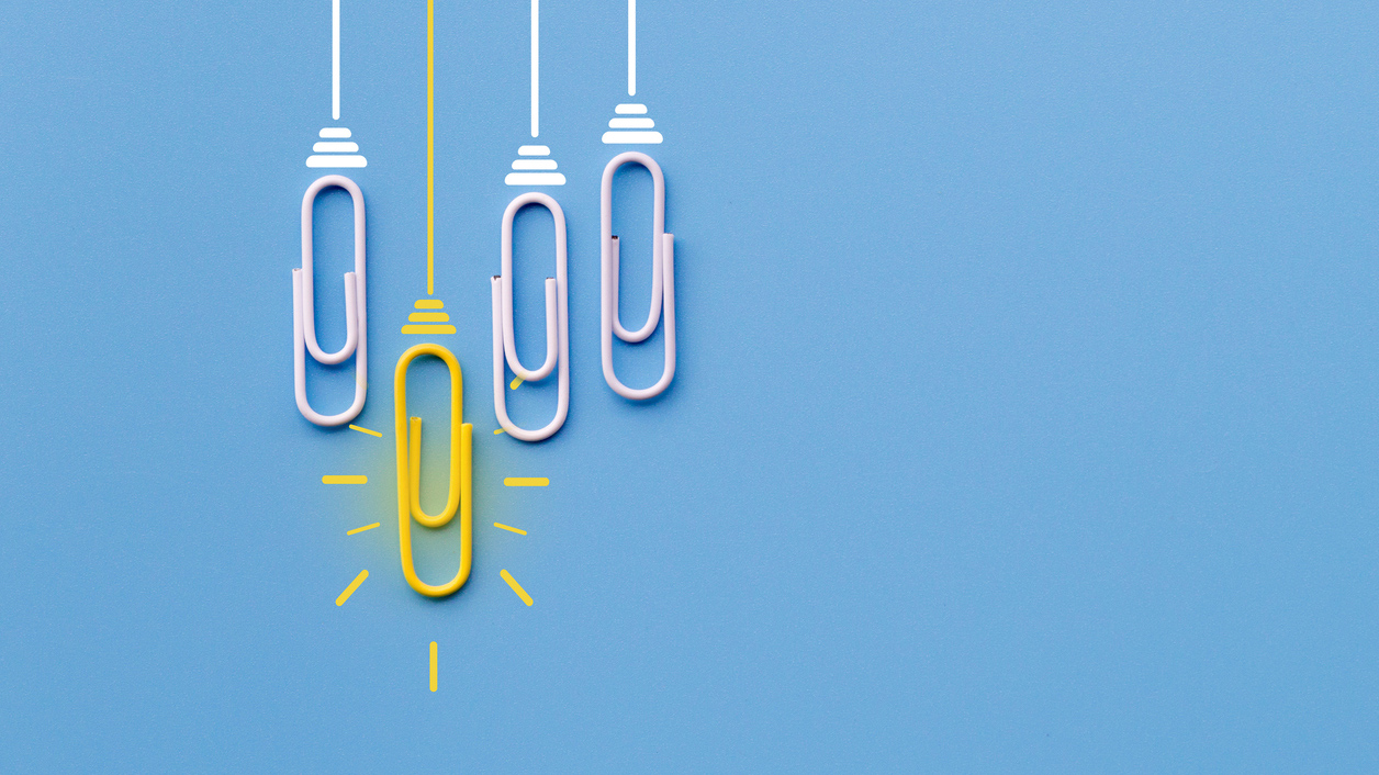 Mehrere Büroklammern hängen wie Lampen vor einem blauen Hintergrund, eine von ihnen leuchtet gelb.