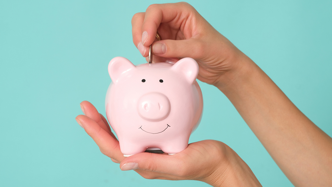 Eine Person hält ein rosa Sparschwein mit freundlichem Gesicht in der Hand und schmeißt mit der anderen eine Münze hinein.