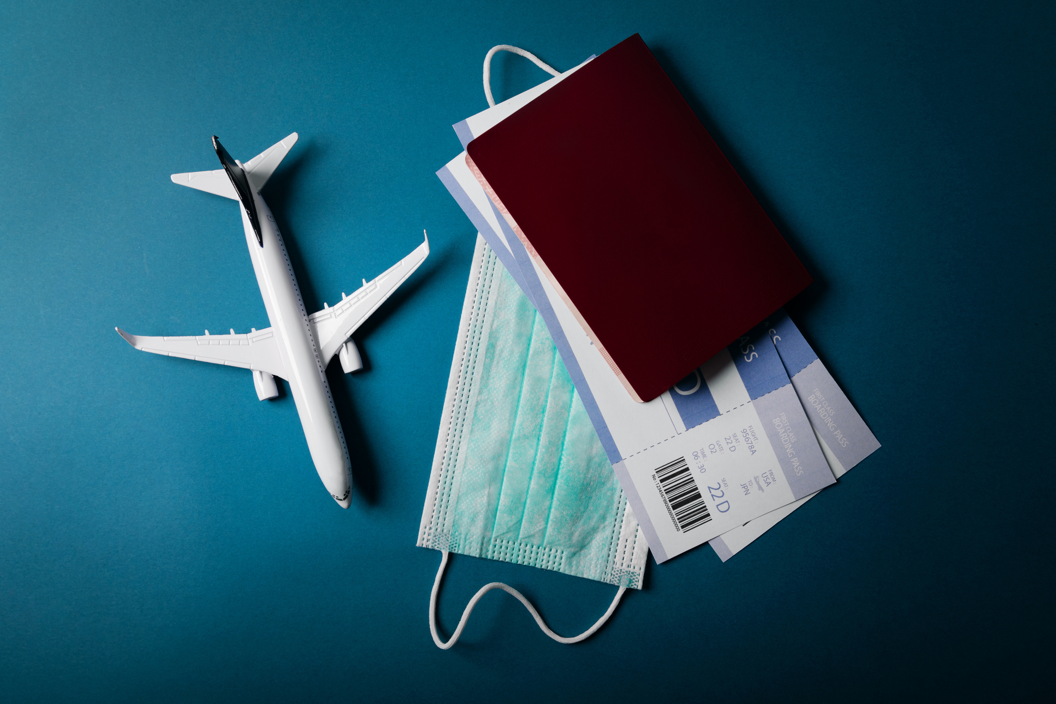 Auf einem petrolfarbenen Hintergrund liegt ein Flugzeug, ein Reisepass, Flugtickets sowie eine medizinische Maske.