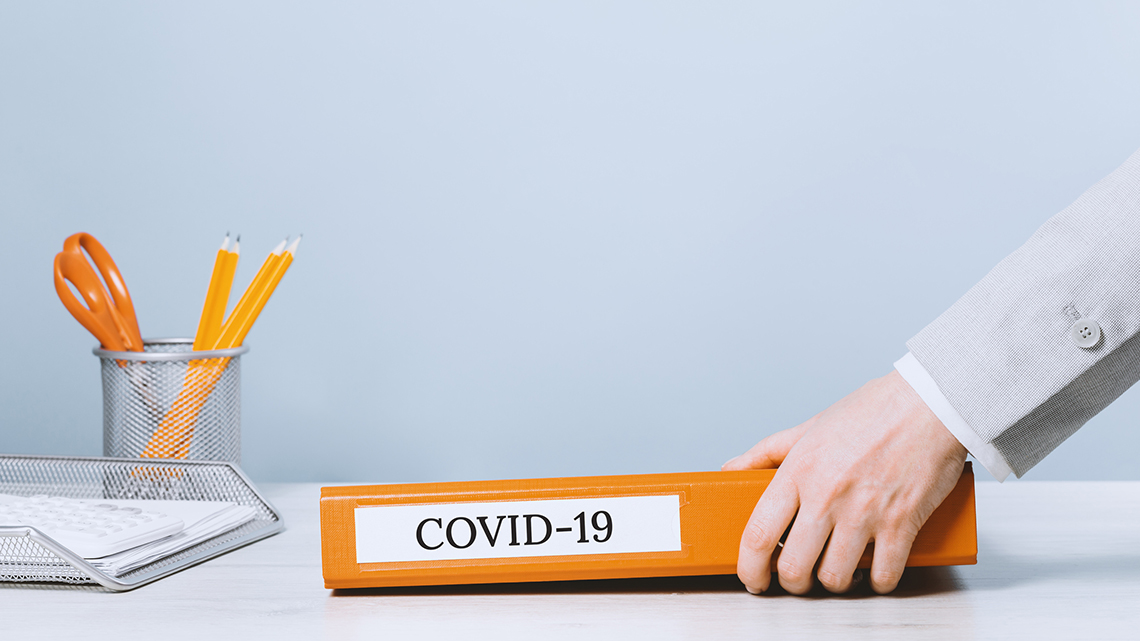 Eine Person legt einen orangenen Ordner mit der Aufschrift "Covid 19" auf einen Schreibtisch.