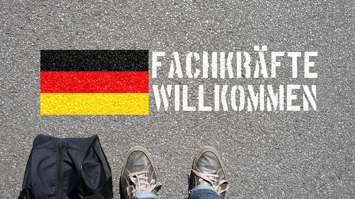 Auf der Straße sind eine Deutschlandfahne und die Worte "Fachkräfte Willkommen" gesprüht. Davor sind die Füße einer Person und eine Reisetasche zu sehen. 