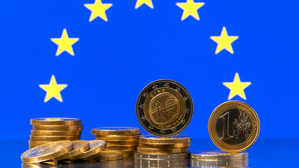 Mehrere Stapel Euromünzen liegen auf einer Glasoberfläche. Im Hintergrund sieht man einen Teil der Europäischen Flagge