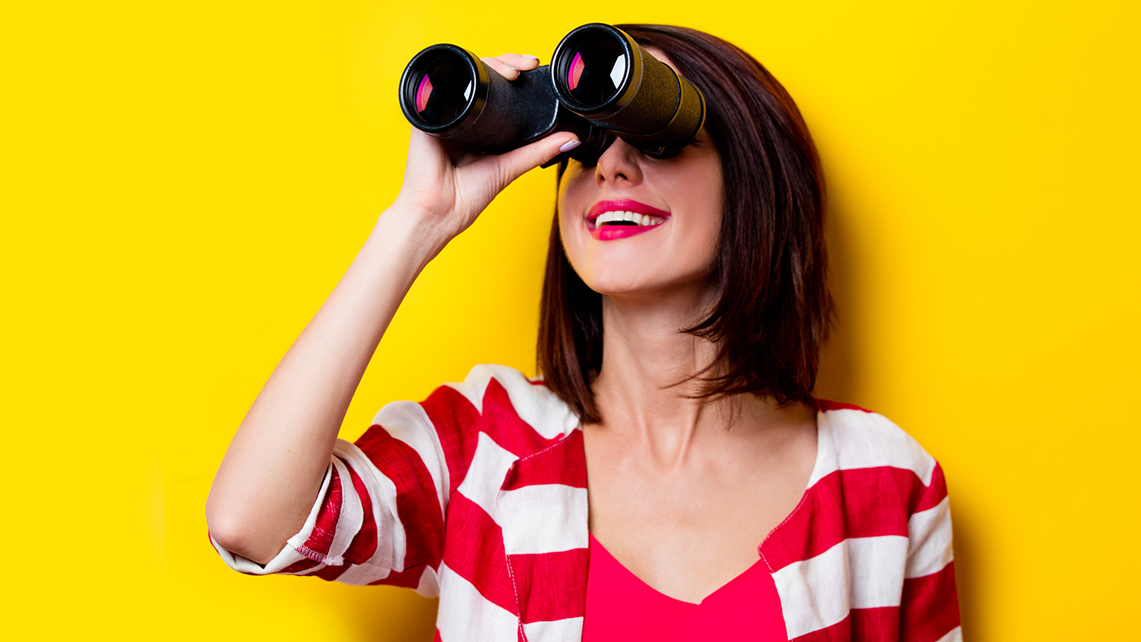 Junge Frau vor gelbem Hintergrund schaut lachend durch ein Fernglas.