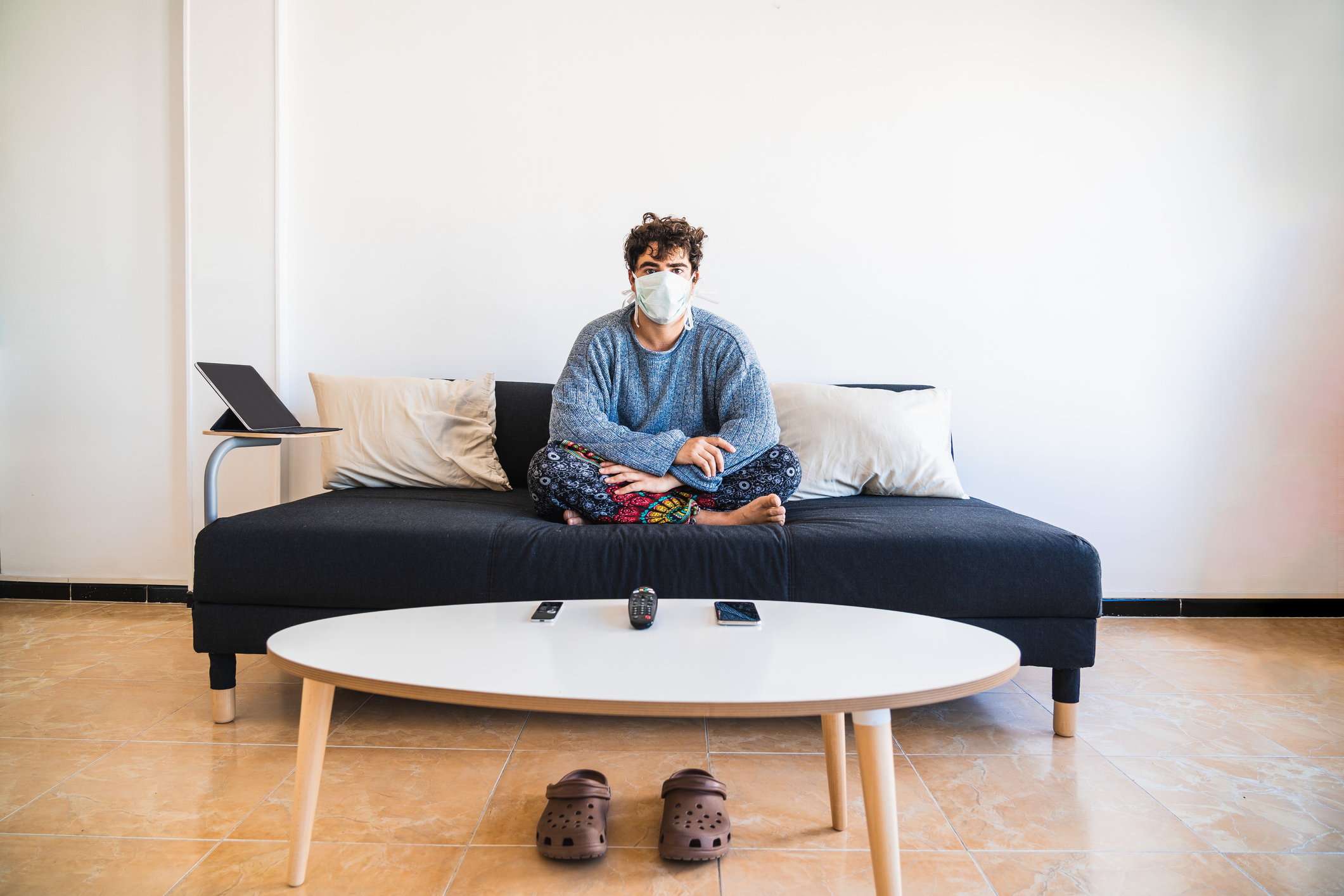 Auf einem Sofa sitzt ein junger Mann in Jogginghose und mit Maske in der Quarantäne. 
