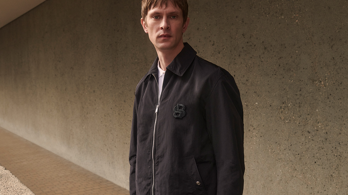 Ein Model trägt eine dunkle Jacke mit dem neuen Hugo Boss Double B Monogramm.