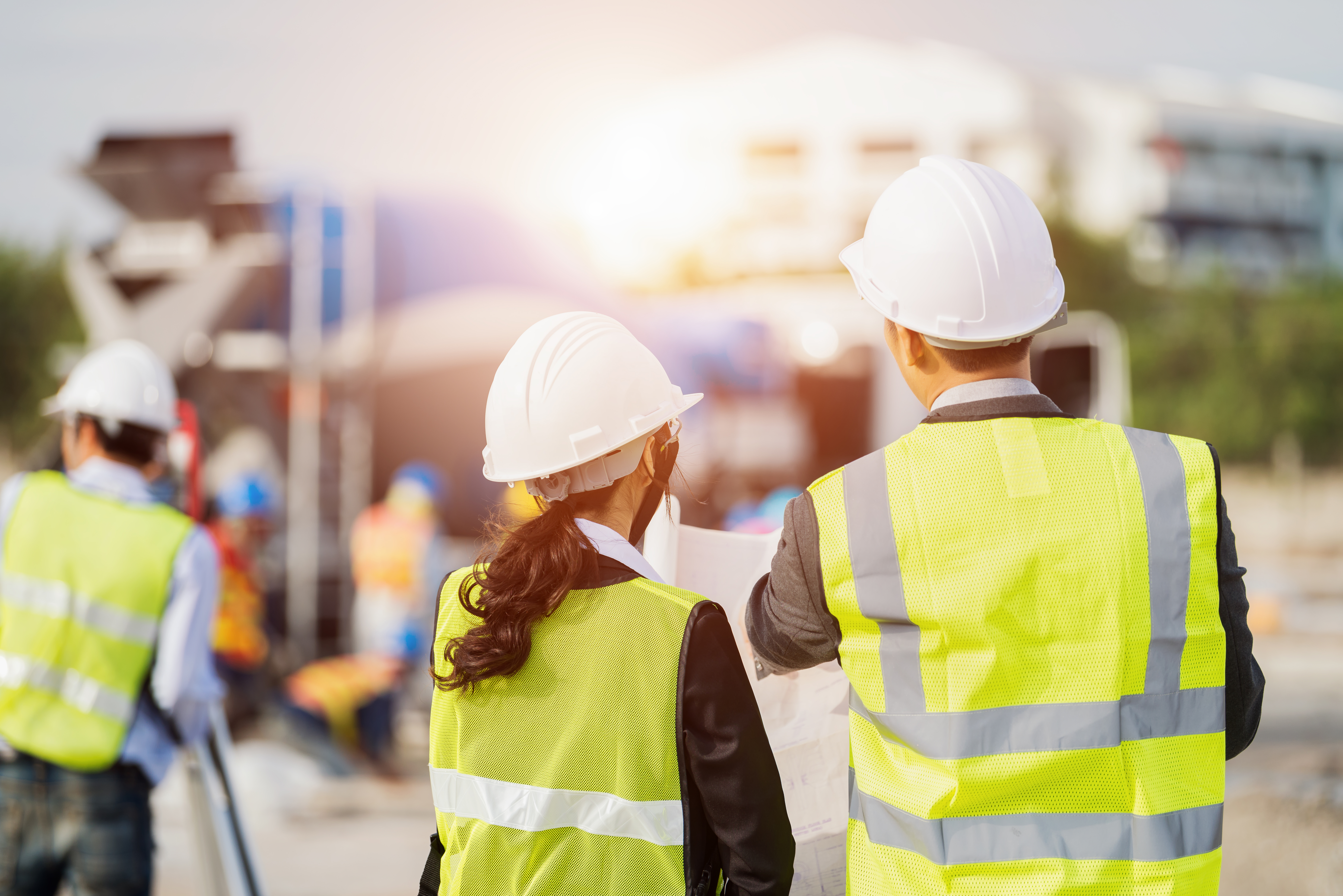 Mann und Frau gehen über Baustelle und tragen dabei Schutzkleidung und Schutzhelm.