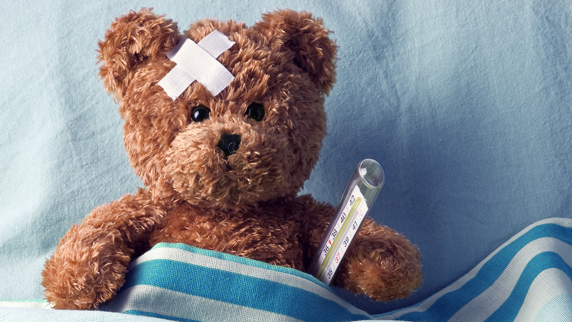 Teddybär mit Thermometer unter dem Arm und mit Pflaster am Kopf liegt zugedeckt auf einem Kissen.