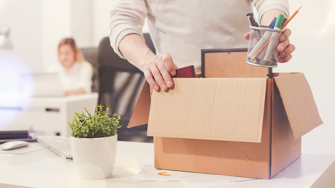 Zwei Hände packen den Inhalt eines Schreibtischs in eine Pappschachtel. Im Hintergrund sitzt eine Frau an ihrem Schreibtisch.