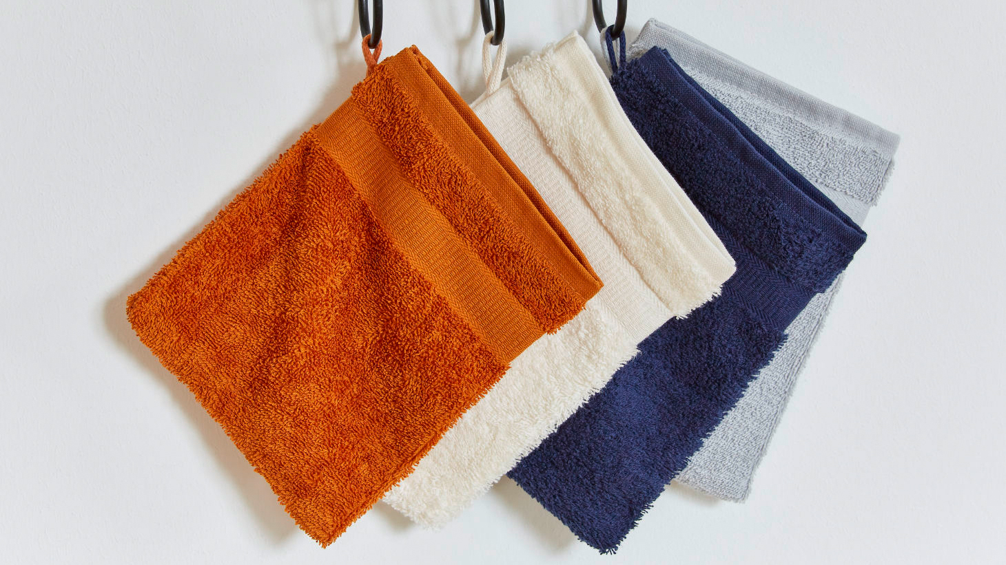 An Haken hängen vier verschiedene Waschlappen in einem dunklen Orange, hellen Beige, dunklen Blau und hellen Blau. 