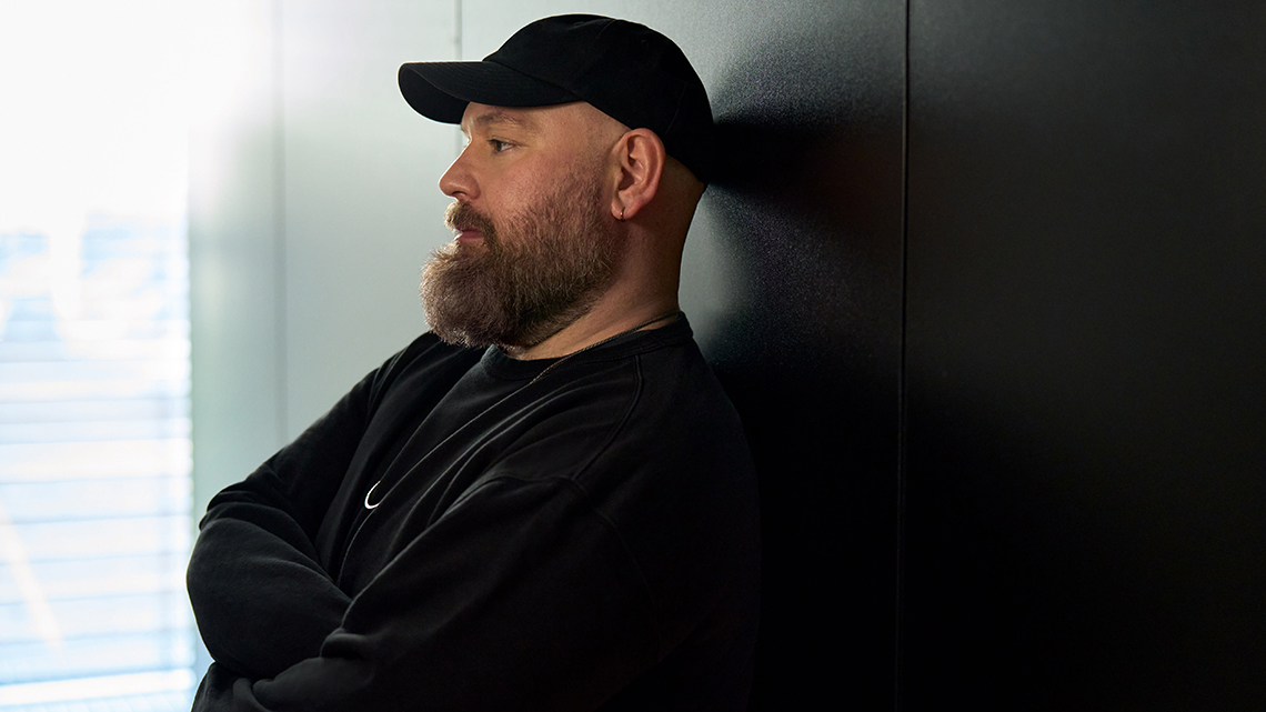 Der neue Creative Director von Hugo Boss, Marco Faliconi, lehnt in einem dunklen Pullover und mit einer dunklen Kappe an einer dunklen Wand.