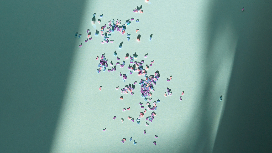 Auf einem mintgrünen Hintergrund der von Sonne und Schatten angeleuchtet ist liegen rosafarbene Krümel Mikroplastik.