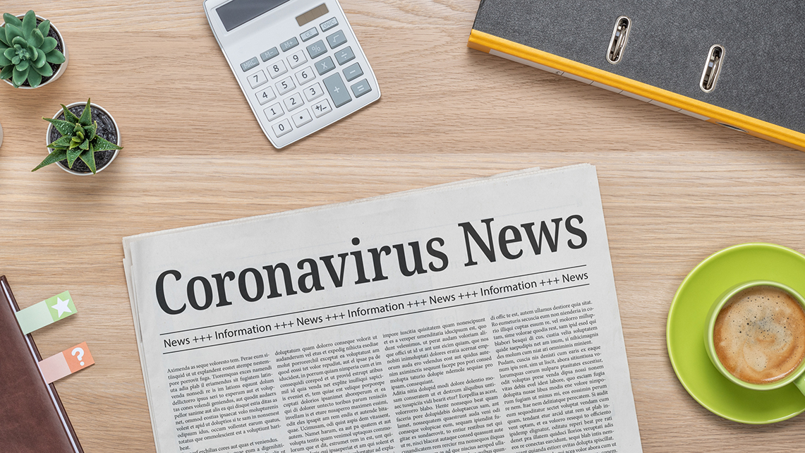 Zeitung mit Überschrift Coronavirus News liegt auf einem Tisch neben einem Ordner, einem Taschenrechner und einer Tasse Kaffee.