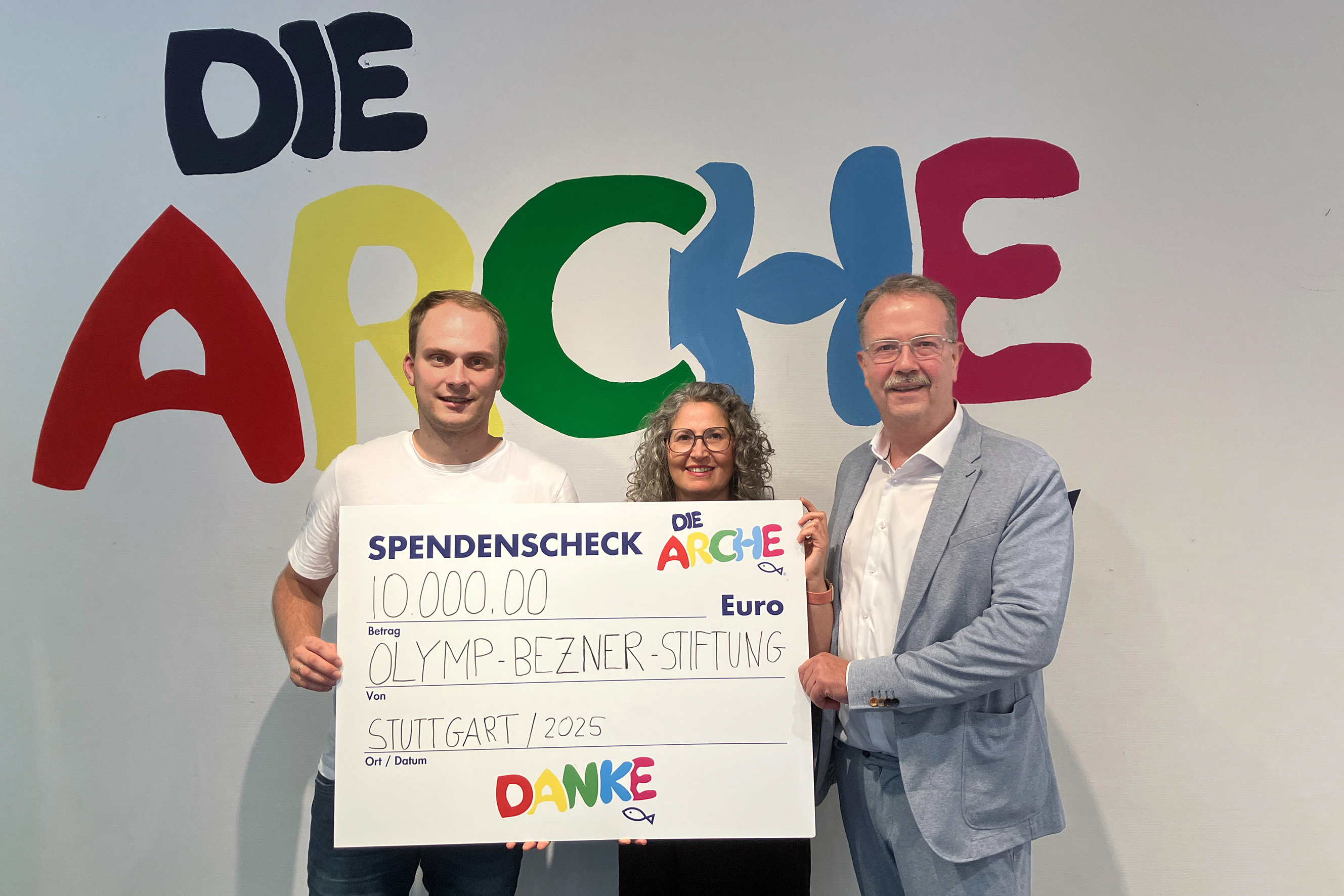Drei Personen bei der Scheckübergabe von einer Wand mit bunter Schrift Die Arche.