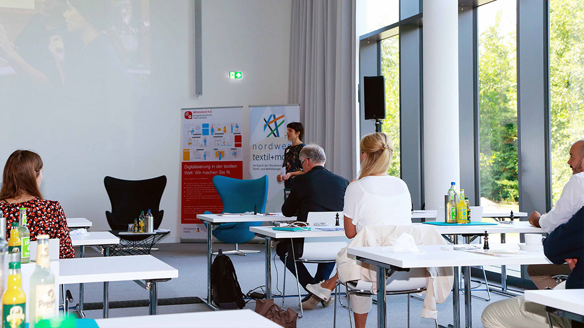 Bei der Textil vernetzt Fachtagung in Münster sieht man Dr. Maria Rost das Kompetenzzentrum vorstellen. Davor sitzen einige Zuhörer ihres Vortrags. 