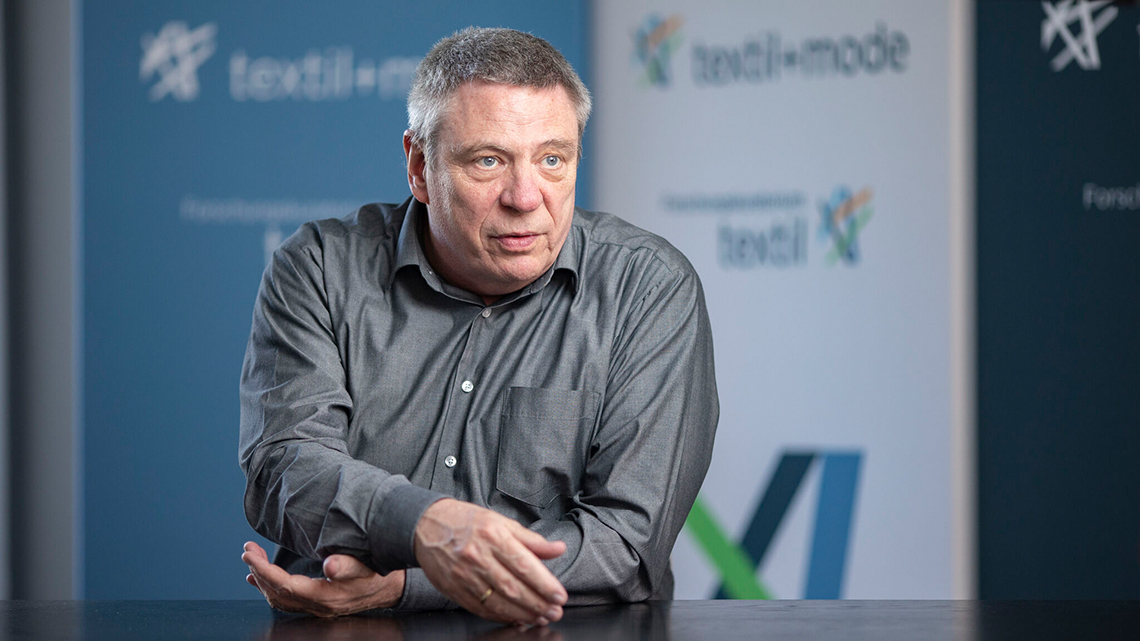 Uwe Mazura, Hauptgeschäftsführer textil+mode, sitzt vor dem Logo des Verbandes und erklärt einen Sachverhalt.