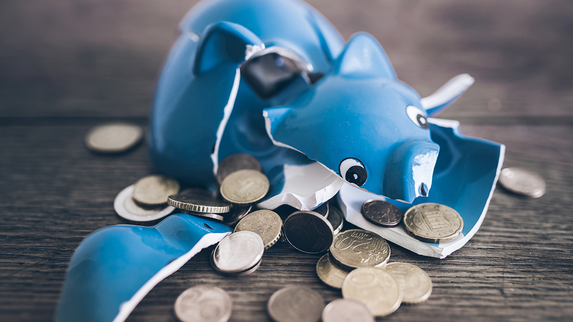 Scherben eines zerbrochenes blaues Sparschwein liegen vermischt mit Münzen auf einem Holztisch