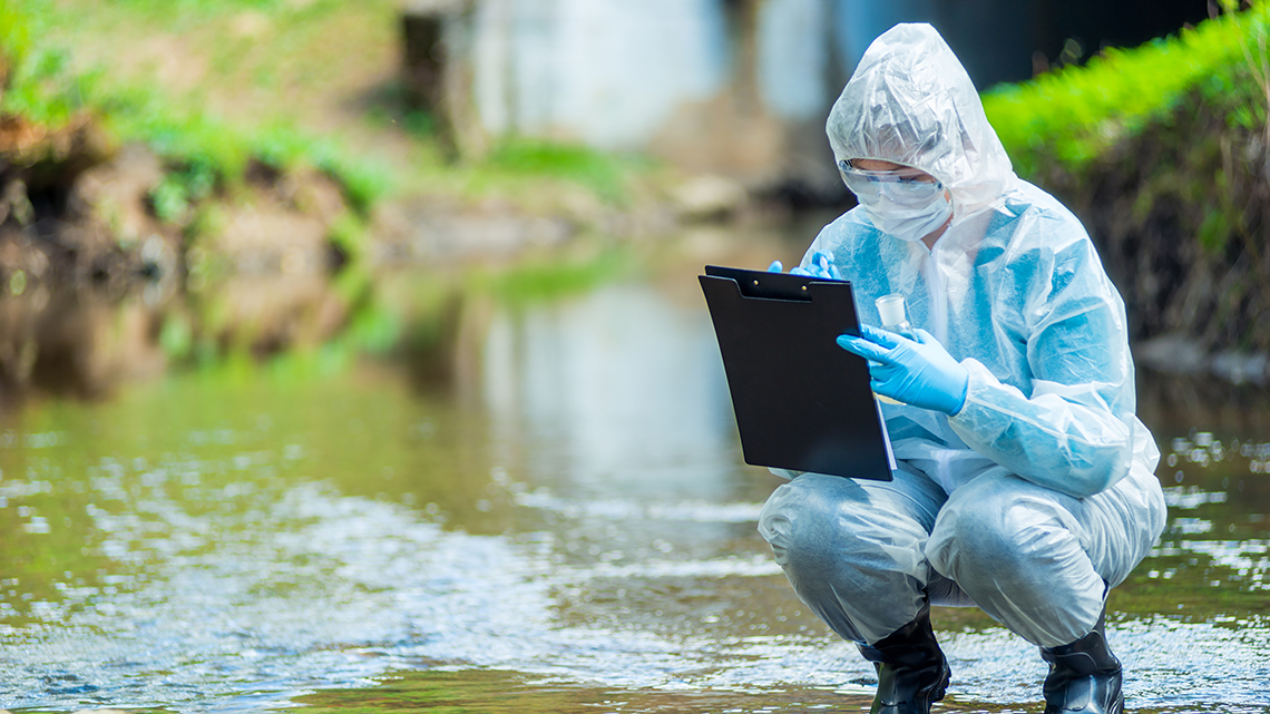 Eine in Schutzkleidung gehüllte Person kniet in einem Flussbett und nimmt Wasserproben. 